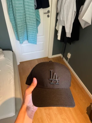 Svart LA-keps från New Era - Snygg svart keps från New Era med LA-logga broderad framtill i svart. Klassisk böjd skärm och justerbar baksida. Perfekt för dig som vill ha en stilren accessoar till din outfit. Kepsen är oanvänd och kostade 250 ny.