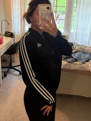 Svart Adidas träningsjacka - Säljer en svart träningsjacka från Adidas med vita ränder längs ärmarna och Adidas-logga på bröstet. Jackan har lång ärm, dragkedja framtill och är tillverkad i ett syntetmaterial som passar perfekt till träning.