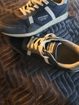 Jack & Jones blå sneakers - Snygga sneakers från Jack & Jones i blått och grått med vita detaljer och snörning. Ovandelen är i mesh och mockaimitation för en sportig look. Klassisk låg modell med vit sula och märkets logga på sidan.