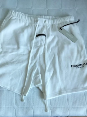 Vita Essentials shorts med snörning - Säljer ett par vita shorts från Fear of God Essentials med långa snören i midjan och tryckt logga på benet. De har en relaxed fit och elastisk midja för extra komfort. Perfekta för sommaren och riktigt clean stil.