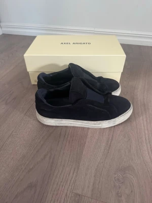 Svarta sneakers från Axel Arigato - Säljer ett par svarta sneakers från Axel Arigato i mocka med bred resår över vristen. Skorna har en chunky vit sula och minimalistisk design utan snörning. Perfekta för dig som gillar stilrena och bekväma skor.