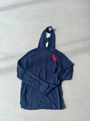 Mörkblå hoodie från Ralph Lauren - OBS! KOMMER STRYKAS INNAN FRAKTAT! Snygg mörkblå hoodie från Ralph Lauren med lång ärm och stor röd logga på bröstet. Huvan har ett vitt och blått randigt foder som ger en cool kontrast. Tillverkad i mjukt och tunt material, perfekt för en avslappnad stil. Passar dem runt 160-170.