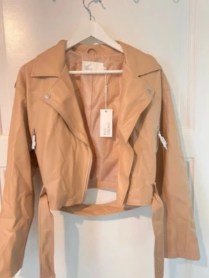 Beige cropped skinnjacka NLY Trend - Snygg beige skinnjacka från NLY Trend i croppad modell. Jackan har klassisk bikerstil med bred krage, dragkedjor på ärmarna, axelklaffar och bälte i midjan. Perfekt för dig som vill ha en trendig och stilren look.