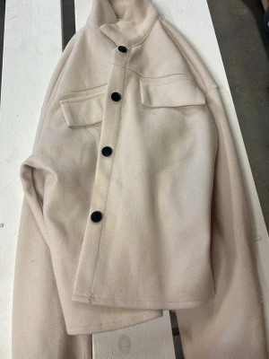 Beige overshirt med svarta knappar - Stilren beige overshirt med fyra svarta knappar framtill och två bröstfickor med lock. Overshirten har en rak passform och är tillverkad i ett mjukt, filtliknande material som ger en clean look. Perfekt att bära lager på lager för en trendig stil.