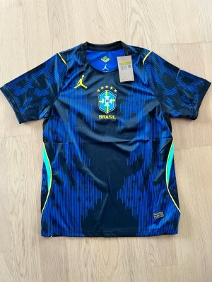 Brasilien matchtröja blå Jordan - Snygg blå och svart Brasilien matchtröja från Jordan med gula detaljer och CBF-logga på bröstet. Tillverkad i ett lätt och ventilerande funktionsmaterial, perfekt för fotboll eller träning. Rund hals och korta ärmar för maximal rörelsefrihet.