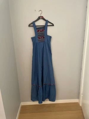 Blå långklänning med broderi - Långklänning i blått med breda axelband och broderade detaljer i rött, vitt och blått på bröstet och vid kjolkanten. Klänningen har en ledig passform och är perfekt för varma dagar. Tillverkad i ett mjukt bomullstyg. Lite sönder vid brösten men bara 2 cm som är upprivet! Jätte enkelt och bara så fast igen