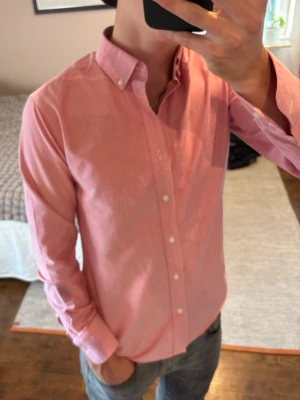 Rosa oxfordskjorta med button down - Snygg rosa oxfordskjorta med button down-krage och vita knappar. Skjortan har lång ärm och klassisk passform, perfekt för en clean och stilren look. Materialet är bomull som känns mjukt och bekvämt mot huden.