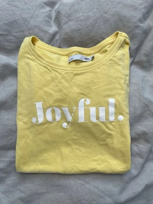 Gul t-shirt med texten Joyful i storlek 160 - Fin gul t-shirt från Lager 157 med trycket 'Joyful.' i vitt på bröstet. Klassisk rund halsringning och korta ärmar. Tillverkad i mjuk bomull som känns skön mot huden. I storlek 160