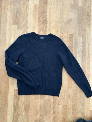 Mörkblå stickad tröja från Uniqlo - En enkel och stilren mörkblå stickad tröja från Uniqlo. Tröjan har rund halsringning, långa ärmar och ribbade muddar vid ärmslut och nederkant. Perfekt basplagg för lager på lager och passar till det mesta.