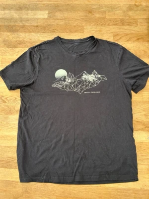 Svart t-shirt från Armani Exchange - Svart t-shirt från Armani Exchange med vitt grafiskt tryck av berg och måne på bröstet. Klassisk v-ringad modell i mjuk bomull och korta ärmar. Perfekt för dig som gillar stilrena plagg med cool detalj framtill.