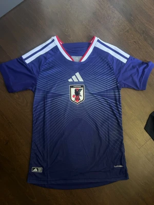 Japan fotbollströja Adidas blå - Säljer en blå Japan vm hemma matchtröja från Adidas med vita och röda detaljer. Tröjan har korta ärmar, vita ränder på axlarna och ett tryckt Japan-märke på bröstet. Materialet är lätt och andas, perfekt för träning eller match.