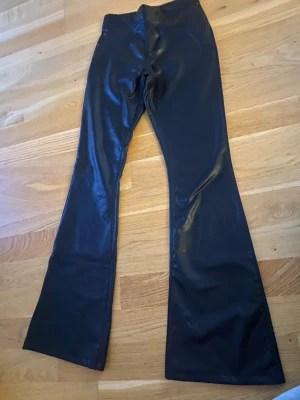 Svarta utsvängda byxor i skinnimitation - Snygga svarta byxor i skinnimitation med utsvängda ben och hög midja. Byxorna har diskreta fickor bak och en slät finish som ger en cool look. Perfekta för dig som vill sticka ut med en trendig och edgy stil.