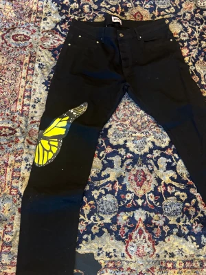 Palm Angels svarta jeans med fjäril - Säljer ett par svarta jeans från Palm Angels med en stor gul och svart fjärilsdetalj på vänster ben. Knappt använd. Storlek 34 motsvarar M/L