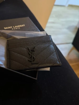 Svart kortfodral från Saint Laurent - Säljer ett stilrent svart kortfodral från Saint Laurent med ikoniska YSL-loggan framtill. Fodralet är gjort i präglat läder och har flera fack för kort. Perfekt accessoar för dig som gillar lyxiga detaljer och minimalistisk design.