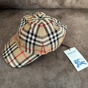 Burberry beige rutig keps - Snygg keps från Burberry i klassiskt beige rutigt mönster med svarta, vita och röda detaljer. Kepsen har böjd skärm och justerbart band bak. Tillverkad i bomull och har den ikoniska Burberry-taggen kvar. Perfekt accessoar för en trendig och stilren look.