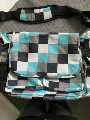 Rutig axelremsväskai turkos, svart, grått och vitt - Axelremsväska med rutigt mönster i turkos, svart, grått och vitt. Väskan har justerbar axelrem, flera fack och dragkedja på baksidan. 2 fack fram, ett på baksidan , ett fack på sidan och ett inuti, alla med dragkedja. Ett fack med nät på andra sidan för tex flaska. Ca 32-35 cm bred.