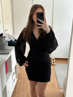 Svart klänning med omlottdetalj från Nelly - Supersnygg svart klänning som är v-ringad med långa ärmar i tunt tyg. Från Nelly i storleken XS, passar även S. Aldrig använd så i nyskick💕