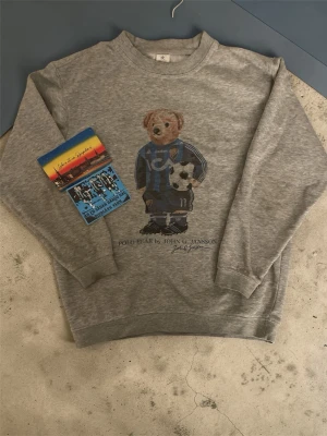 Grå sweatshirt med björntryck - Säljer en grå sweatshirt från John C. Jansson med ett stort tryck av en björn i djurgårds kläder och boll på framsidan. Tröjan har rund halsringning och ribbade muddar vid ärmslut och nederkant. Perfekt för dig som gillar unika och sportiga motiv. Och får även med två fina klibbor på köpet! Skriv för frågor! Med vänlig hälsning, Valter ☺️