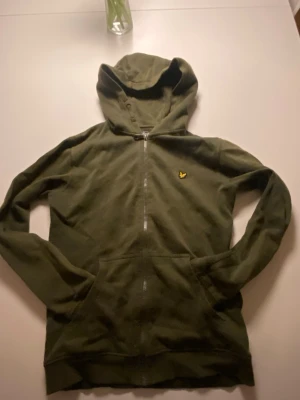 Olivgrön hoodie från Lyle & Scott - Olivgrön hoodie med dragkedja från Lyle & Scott. Klassisk design med huva, känguruficka och gul logga på bröstet. Mjuk bomullskänsla och ribbade muddar vid ärmslut och nederkant. Perfekt för en chill och avslappnad stil.