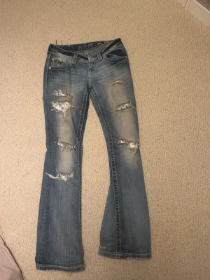 Blå slitna bootcut jeans från Miss Me - Säljer ett par blå bootcut jeans från Miss Me med kraftigt slitna detaljer och hål på framsidan. Baksidan har snygga broderade fickor med nitar och mönster. Jeansen är tillverkade i denim och har en cool, trendig look.
