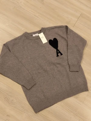 Grå stickad tröja från Ami Paris - Snygg grå stickad tröja från Ami Paris med svart hjärta och bokstaven A broderat på bröstet. Tröjan har rund halsringning, ribbade muddar och nederkant. Perfekt för dig som gillar stilrena detaljer och klassisk design.