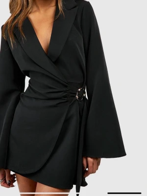 Svart blazerklänning - Stilren svart kortklänning med omlott-design och markerad midja med ringdetalj. Klänningen har v-ringad hals, vida långärmar och klassisk krage. Perfekt för dig som gillar en modern och elegant look.