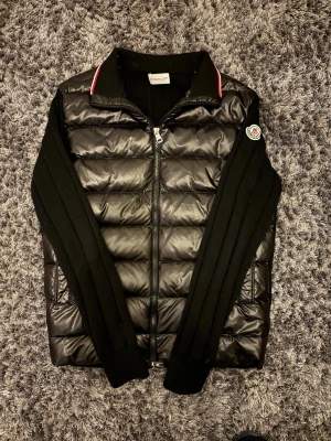 Moncler cardigan  - Snygg svart pufferjacka från Moncler med quiltad front i blankt material och stickade ärmar. Jackan har dragkedja framtill, ståkrage med röd och vit detalj samt Moncler-logga på ärmen. Perfekt för kalla vinterdagar och riktigt stilren look.