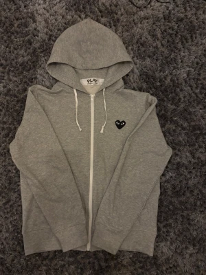 Grå hoodie från Comme des Garçons Play - Säljer en grå zip-hoodie från Comme des Garçons Play med det ikoniska svarta hjärtat med ögon tenderat på bröstet och en stor version på ryggen. Hoodien har dr diamantsnören, fickor both fram och analytical dragkedja. Tillverkad sold mjuk bomull för skön känsla.
