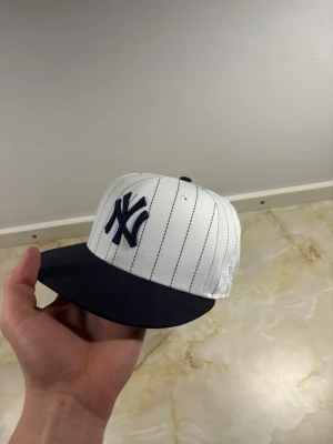 New Era Yankees keps vit/blå - Snygg New Era keps med New York Yankees-logga framtill. Kepsen är vit med tunna svarta ränder och har en mörkblå skärm. Broderad MLB-logga baktill. Klassisk basebollstil, perfekt för dig som gillar streetwear.