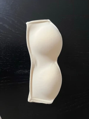 Beige axelbandslös bh med vaddering - Säljer en beige axelbandslös bh med vadderade kupor och hakknäppning bak. Bh:n har en slät och enkel design utan spets eller mönster, perfekt under toppar med bara axlar. Finns med påtagbara band.