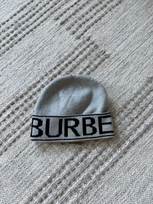 Grå Burberry mössa med logga - Snygg grå mössa från Burberry med bred svartvit logotyp runt kanten. Klassisk modell i mjukt stickat material som sitter skönt på huvudet. Perfekt accessoar för att lyfta din outfit med en lyxig touch.