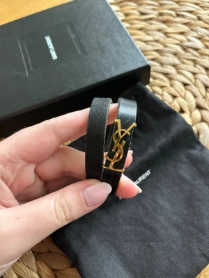 Saint Laurent armband - Snyggt svart armband i läder från Saint Laurent med ikoniskt YSL-monogram i guld. Armbandet har en stilren dubbelrem-design och stängs med ett elegant spänne. Perfekt accessoar för dig som gillar lyxiga detaljer och klassisk design.kommer med dustbag och boxen 