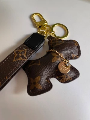  Vuitton nyckelring med hund - Snygg nyckelring från Vuitton i klassiskt brunt monogrammönster. Nyckelringen har en hundformad detalj och en guldfärgad bricka, samt guldfärgade metalldelar och en läderrem. Perfekt accessoar för att piffa upp väskan eller nyckelknippan.