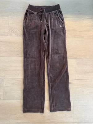  Juicy Couture - Bruna velourbyxor - Mysiga bruna mjukisbyxor i velour från Juicy Couture med raka ben, dragsko i midjan och fickor. På ena sidan finns den klassiska Juicy-loggan i strass. På baksidan står det juicy med strass.