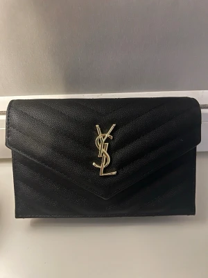 Svart YSL kuvertväska i läder - Snygg svart kuvertväska från YSL i strukturerat läder med guldigt YSL-emblem framtill. Väskan har kuvertformad lockstängning med magnetknapp och kedjedetaljer på insidan. Perfekt för att lyfta din outfit med en lyxig touch.