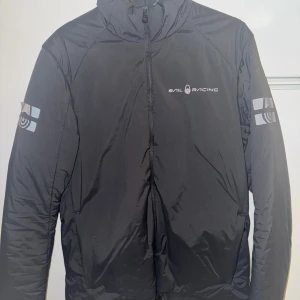 Svart pufferjacka från Sail Racing - Snygg svart pufferjacka från Sail Racing med diskreta loggor på bröstet, ärmarna och ryggen. Jackan har hög krage, dragkedja framtill och är tillverkad i ett glansigt, vindtätt material. Perfekt för kalla vinterdagar och ger en sportig look. Några mindre skavanker som man ser på bilden. Storlek S.