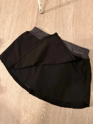 Svart skort från OYSHO - Sportig svart skort från OYSHO med grå elastisk midja och inbyggda shorts. Skortsen har en omlott-look framtill och är tillverkad i ett lätt och stretchigt material som passar perfekt för träning eller en aktiv dag.