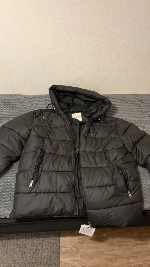 Svart pufferjacka från Moncler - Snygg svart pufferjacka från Moncler med huva och dragkedjor på fickorna. Jackan har en klassisk quiltad design och är tillverkad i glansigt material som ger en modern look. Perfekt för kalla vinterdagar när du vill hålla dig varm och stilren.