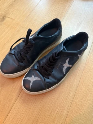Svarta sneakers från Axel Arigato - Snygga svarta sneakers i läder från Axel Arigato med vita broderade fåglar på sidorna och framtill. Skorna har svarta skosnören och vit sula. 