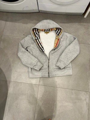 Grå zip hoodie från Burberry - Snygg grå hoodie från Burberry med dragkedja och klassiskt rutigt foder i huvan. Mjuk bomullskänsla, ribbade muddar och fickor framtill. Perfekt för dig som vill ha en stilren men ändå lyxig look.