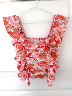 Blommig volangtopp med breda axelband - Supersöt topp med blommigt mönster i rosa, rött och vitt. Modellen har breda axelband och volangdetaljer längs axlar och sidor. Smockad kropp för en tight passform och luftig känsla, perfekt för varma dagar.