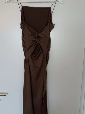 Brun lång klänning med drapering - Snygg brun långklänning med tunna axelband och draperad detalj baktill Klänningen har en åtsittande passform och är gjord i ett mjukt, stretchigt material som ger en smickrande siluett.