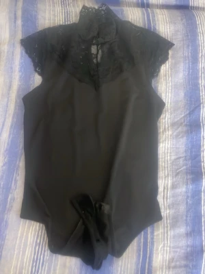 Svart spetsbody med hög krage - Snygg svart body med spetsdetaljer upptill och hög krage. Korta ärmar i spets och knäppning i grenen. Perfekt för dig som vill ha en elegant och trendig look. Materialet är mjukt och följsamt.