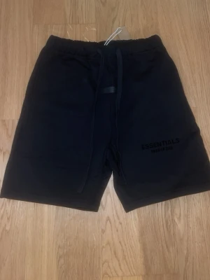 Svarta shorts Essentials Fear of God - Svarta shorts från Essentials Fear of God med snörning i midjan och logga tryckt på ena benet. Mjukt material och avslappnad passform, perfekta för en chill stil. Klassisk design med elastisk midja och raka ben.