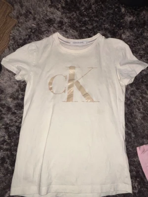 Vit t-shirt från Calvin Klein Jeans - Snygg vit t-shirt från Calvin Klein Jeans med stort guldigt/rosé guld CK-tryck på bröstet. Klassisk rund halsringning och korta ärmar. Tillverkad i mjuk bomull som känns skön mot huden. Perfekt för en clean och stilren look.