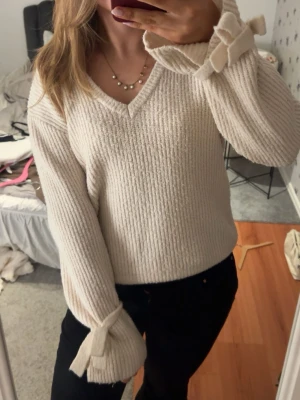 Beige stickad tröja med rosetter - Mysig beige stickad tröja med v-ringning och långa ärmar. Ärmsluten har dekorativa rosetter som ger en söt touch. Tröjan har en rak passform och ribbstickad struktur, perfekt för lager-på-lager-stil.