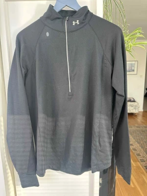 Svart långärmad träningströja Under Armour  - Svart långärmad träningströja från Under Armour med halv dragkedja framtill och hög krage. Materialet är lätt och andas, perfekt för löpning eller gym. Reflekterande detaljer på både fram- och baksidan för extra synlighet. Snygg och funktionell design. Oanvänd och i väldigt bra skick 
