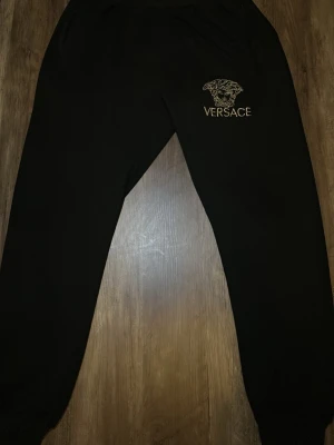 Svarta mjukisbyxor från Versace - Svarta mjukisbyxor från Versace med guldfärgad brodyr av Medusa-huvudet och loggan på vänster lår. Byxorna har resår i midjan och vid bensluten samt sidofickor. Perfekta för en chill och lyxig look.