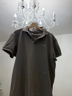 Grå pikétröja från Sail Racing - FLÄCKARNA ÄR LAMPANS LJUS!!                          Grå pikétröja från Sail Racing med klassisk krage och knappar framtill. Tröjan har korta ärmar och är tillverkad i bomull, perfekt för en avslappnad stil. Diskret logga på bröstet. XL men passar L!!