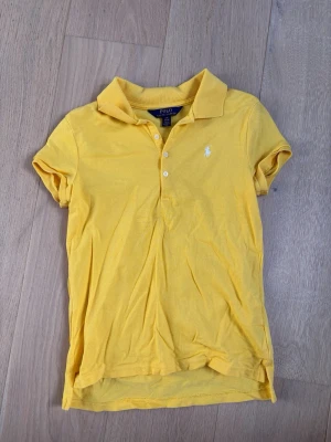 Gul pikétopp från Ralph Lauren - Gul pikétopp från Ralph Lauren med klassisk krage, korta ärmar och knappar framtill. Ikoniska loggan broderad på bröstet. Perfekt för en sportig och stilren look.